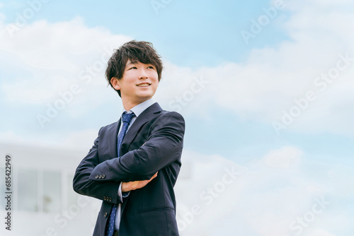 青空とオフィスの前に立つ若いアジア人ビジネスマン（腕組み・見上げる） 