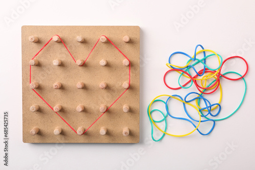 Wooden geoboard with heart made of rubber bands on white table, flat lay. Educational toy for motor skills development photo