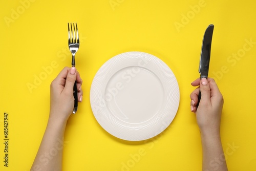 Woman holding fork and knife near empty plate at yellow table, top view