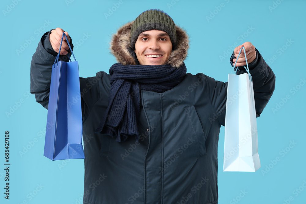 Young man in stylish puffer jacket and scarf with shopping bags on blue background