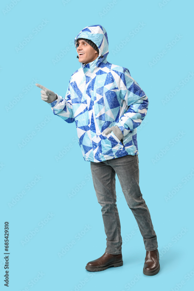 Young man in stylish puffer jacket and gloves pointing at something on blue background Young man in stylish puffer jacket and gloves pointing at something on blue background