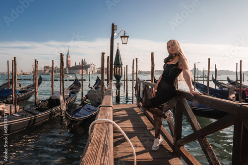A woman in a black dress poses on a pier in Venice, Italy, with gondolas in the background. The serene atmosphere and picturesque scene capture the essence of a summer day in Venice. © Aerial Film Studio