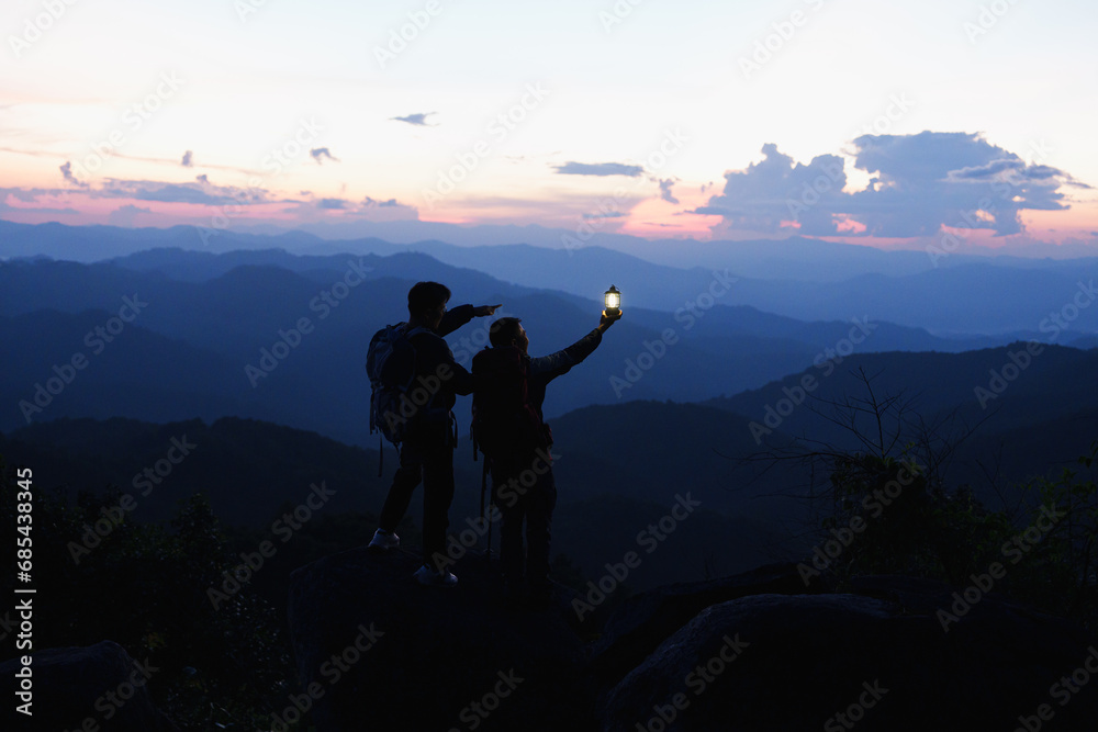 Fototapeta premium Silhouette of Asian two male standing raised hands with trekking poles and kerosene black lamp on cliff edge on top of rock mountain with at sunset rays over the clouds background,