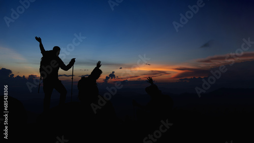 Silhouette of Asian teamwork standing raised hands with trekking poles and Backpack and other camping gear on cliff edge on top of rock mountain with at sunset rays over the clouds background, 