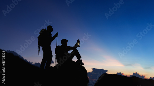 Silhouette of Asian two male standing raised hands with trekking poles and kerosene black lamp on cliff edge on top of rock mountain with at sunset rays over the clouds background,