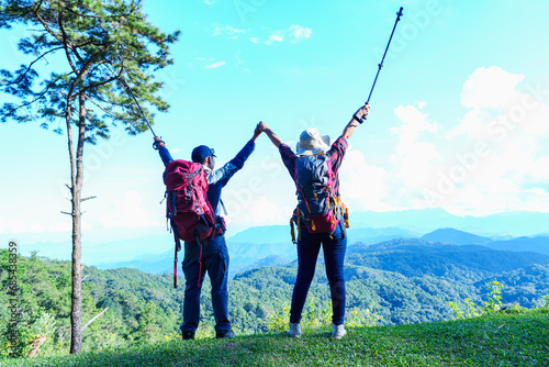 Asian Male and female standing raised hands with trekking poles on Lawn or green grass ground of camping ground, Asia couple hiking in clouds and morning sky background.