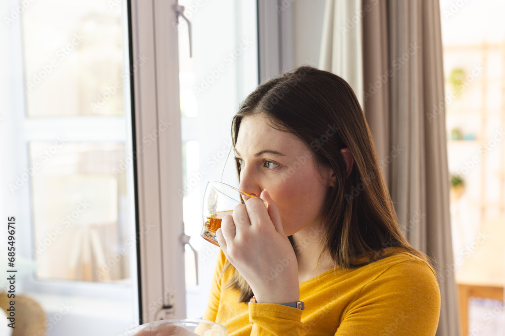 Thoughtful caucasian woman drinking tea by window in living room at home, copy space Thoughtful caucasian woman drinking tea by window in living room at home, copy space