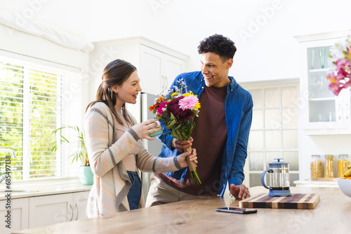 Happy diverse couple holding flowers and holding tea cup in kitchen at home, copy space