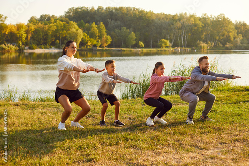 Family stretching after sport. Young family with two children is playing sports outside on bright sunny day. Mom, dad, son, daughter are squatting with outstretched arms against background of lake. photo