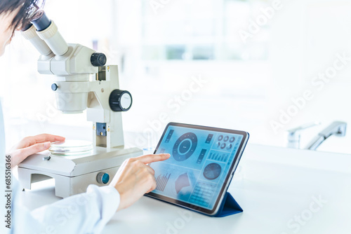 A woman in a white coat researching using a microscope and a tablet.