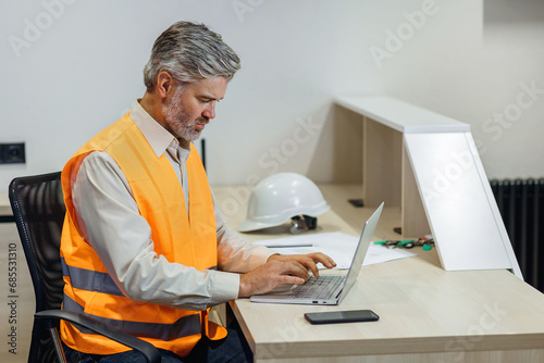 gray haired attractive construction bearded man in vest working with laptop, work on Home building project photo