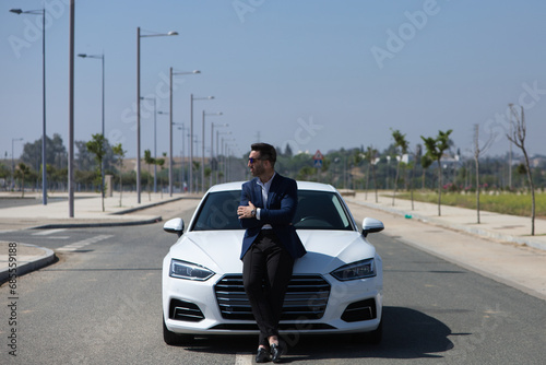 Handsome young businessman with beard and blue suit sitting on his white sports car with his arms folded. The man is wearing sunglasses. Business travel concept. © @skuder_photographer