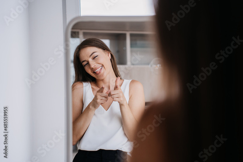 Cheerful young blonde girl smiling at mirror pointing at herself shows tongue being in playful mood ready for ;arty with friends. Careless female, freedom concept. photo