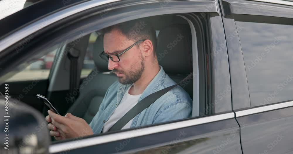 Young Caucasian man sits in a car on a parking lot and typing message on a smart phone
