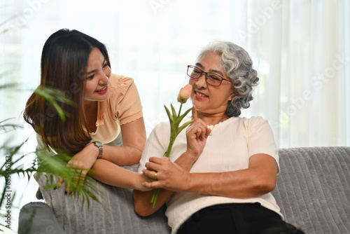 Adult daughter standing behind couch and giving flowers to older mother. Family bonding concept