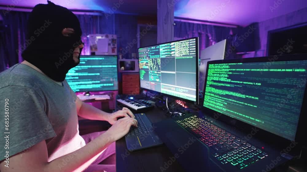 Hacker coding and listening music on programming computer at table. Male anonymous individual looking at notebook and hacking pc in neon color light. IT cybercrime of thief mask hacks virtual database