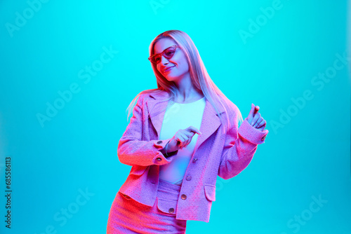Portrait of young blonde woman in pink smart casual costume and style sunglasses against gradient blue background in neon light, filter.