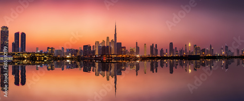 Wide panoramic view of the illuminated skyline of Dubai Business bay with reflections of the modern skyscrapers in the water during dusk time, UAE