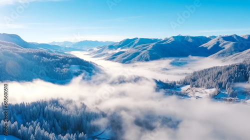 Aerial view of winter mountain valley with fog surrounded by snow-covered forested hills under blue sky