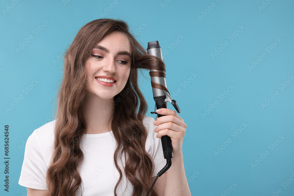 Beautiful young woman using curling hair iron on light blue background, space for text Beautiful young woman using curling hair iron on light blue background, space for text