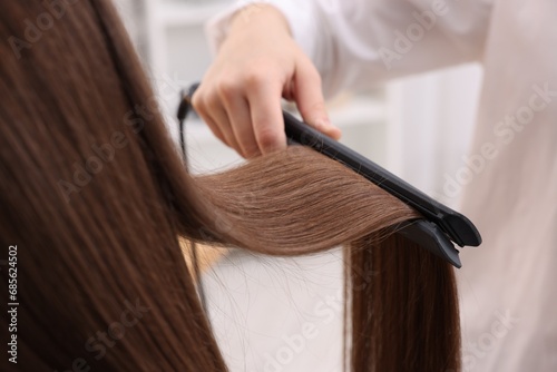 Hairdresser straightening woman's hair with flat iron indoors, closeup