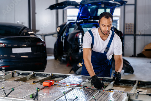 Arabian male technician engages with an EV Car Battery Cell Module troubleshooting a modern electric vehicle battery removed from the vehicle. Mechanic examining a car engine with voltmeter. photo