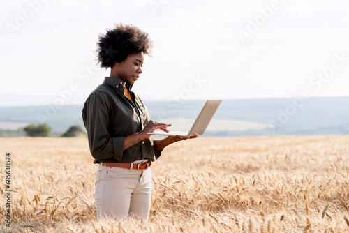 Afro farm worker using laptop amidst barley crops in field photo