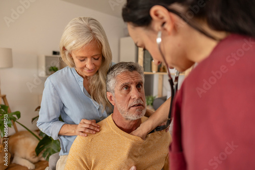 Doctor listening heartbeat of man by woman at home photo