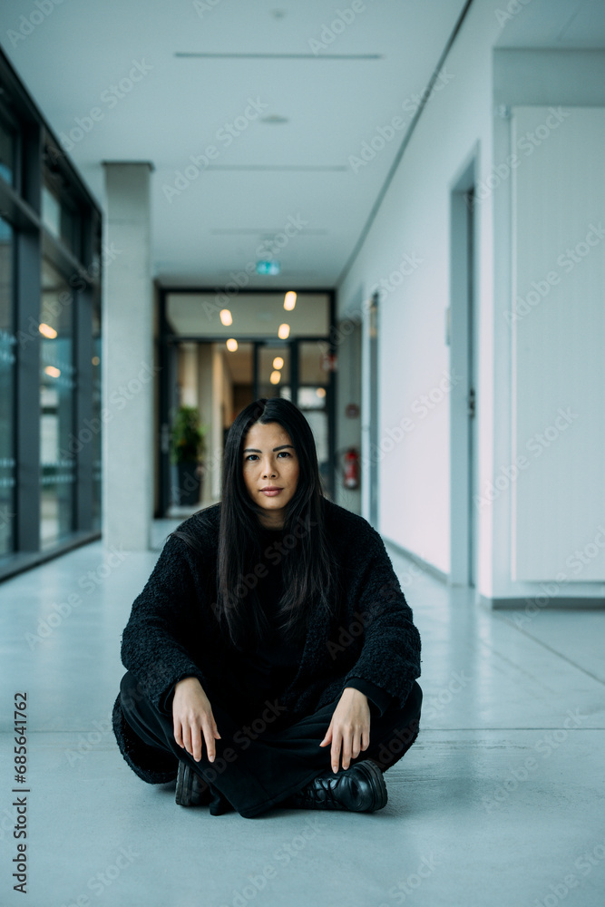 Confident businesswoman sitting cross-legged on floor in corridor at workplace Confident businesswoman sitting cross-legged on floor in corridor at workplace