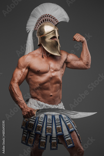 Portrait of an ancient Greek hoplite, posing in a studio with a bare muscular torso, wearing a helmet, and holding a gladius, set against a gray background photo