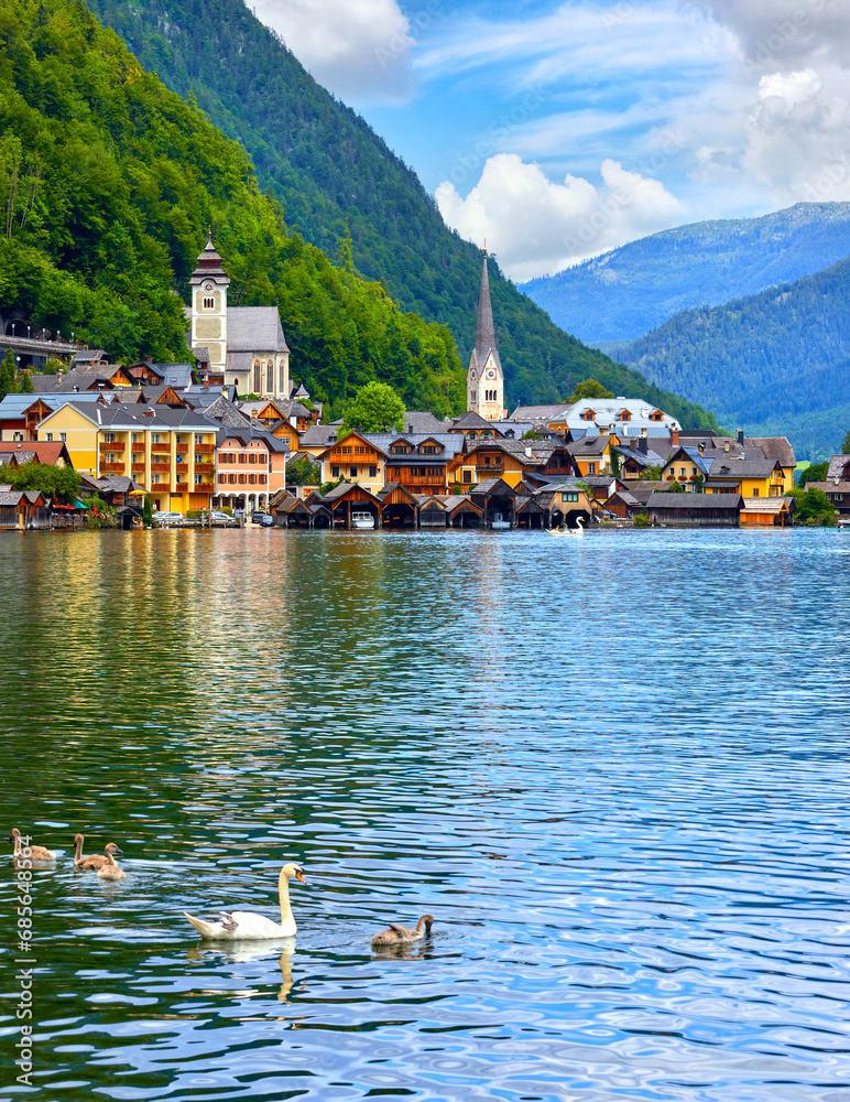 Hallstatt, Austria. View at Hallstattersee Lake and Alps mountains summits. White swan birds near the dock. Ancient houses lake banks with chapel. Summer day. Blue sky clouds
