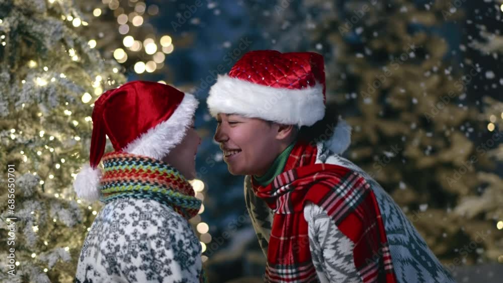 Happy mother with child in warm ornamented sweaters and scarfs playing clappers outdoors, joyfully touching noses. Blurred Christmas trees with garlands. High quality 4k footage