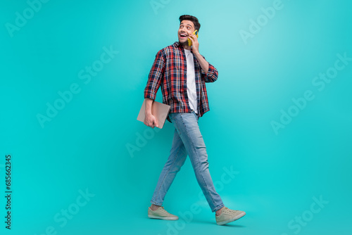 Full length photo of excited dreamy guy wear checkered shirt walking communicating modern device empty space isolated teal color background © deagreez