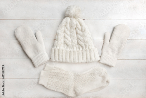 A white knitted hat with a pompom, woolen mittens and socks lie on a white wooden background. Details of winter clothing