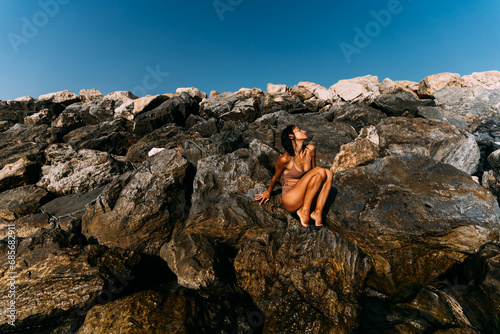 A beautiful, sexy brunette woman is sitting on the rocks in a swimsuit by the sea. © sutulastock