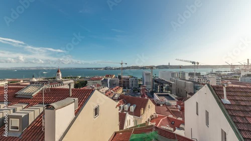Panorama showing red roofs timelapse and 25 de Abril Bridge, Iconic suspension bridge over Tagus River in Lisbon, Portugal. Aerial view from Miradouro de Santa Catarina. Classic Viewpoint at sunny day photo