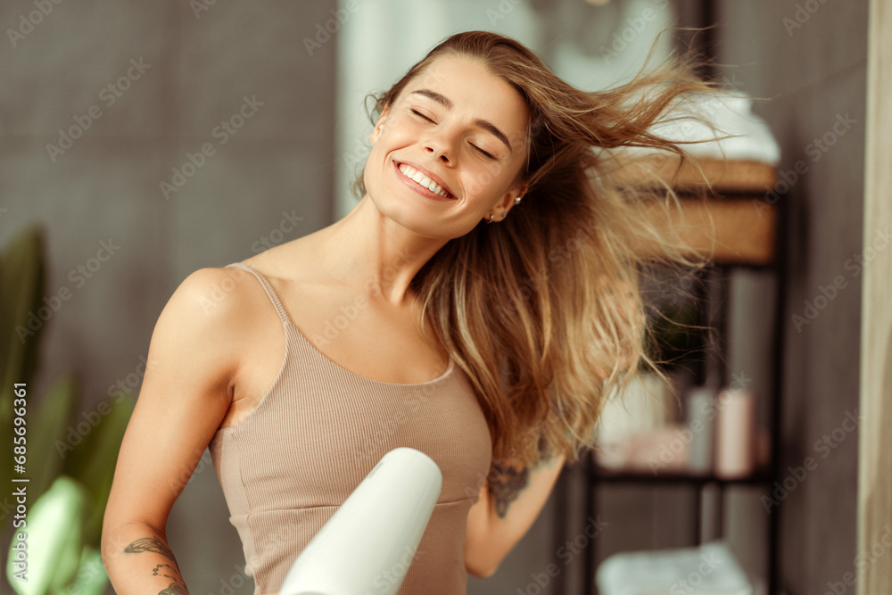 Happy beautiful young woman drying her hair with dryer, wearing tank top standing in bathroom Happy beautiful young woman drying her hair with dryer, wearing tank top standing in bathroom