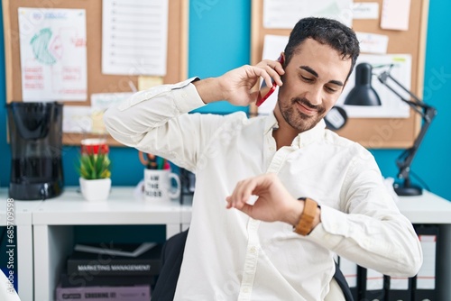 Young hispanic man business worker talking on smartphone looking watch at office