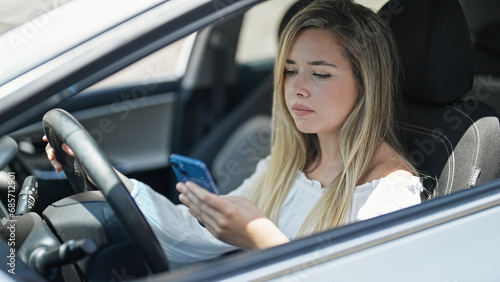 Young blonde woman using smartphone sitting on car at street