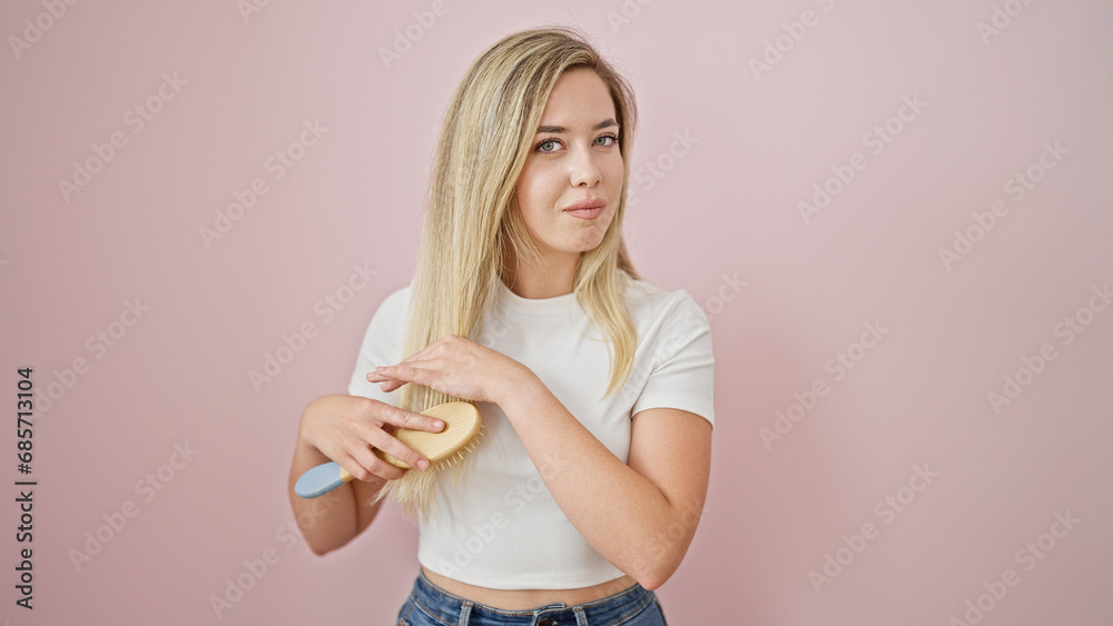 Young blonde woman combing hair smiling over isolated pink background Young blonde woman combing hair smiling over isolated pink background