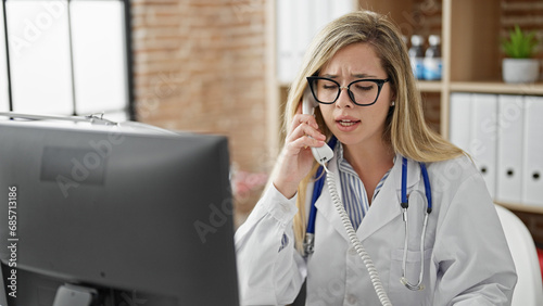 Young blonde woman doctor using computer talking on telephone at clinic