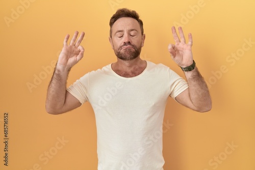 Middle age man with beard standing over yellow background relaxed and smiling with eyes closed doing meditation gesture with fingers. yoga concept.