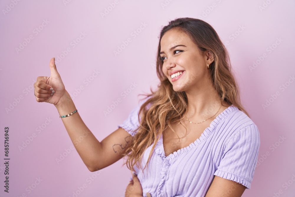 Young hispanic woman standing over pink background looking proud, smiling doing thumbs up gesture to the side Young hispanic woman standing over pink background looking proud, smiling doing thumbs up gesture to the side