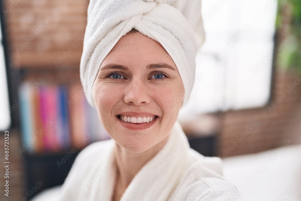 Young blonde woman wearing bathrobe sitting on bed smiling at bedroom Young blonde woman wearing bathrobe sitting on bed smiling at bedroom