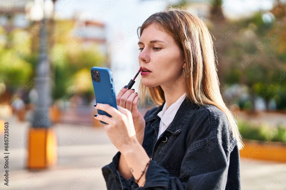 Young blonde woman using smartphone as a mirror make up lips at park Young blonde woman using smartphone as a mirror make up lips at park