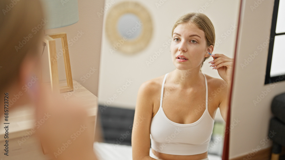 Young blonde woman cleaning ear with cotton stick pad looking on mirror at home Young blonde woman cleaning ear with cotton stick pad looking on mirror at home