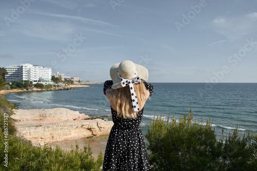 Young woman in hat and polka dot dress looks at sea. Back view. Salou, Costa Dorada in province of Tarragona. Spain.