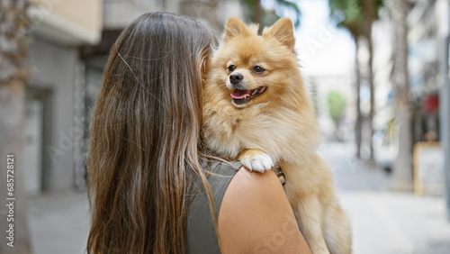 Young hispanic woman, captured from the back standing on a sunny city street, while backwards hugging her beautiful dog, embodying urban pet love and portrait of outdoor expression.