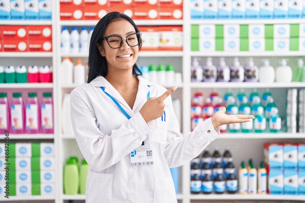 Hispanic woman working at pharmacy drugstore amazed and smiling to the camera while presenting with hand and pointing with finger.