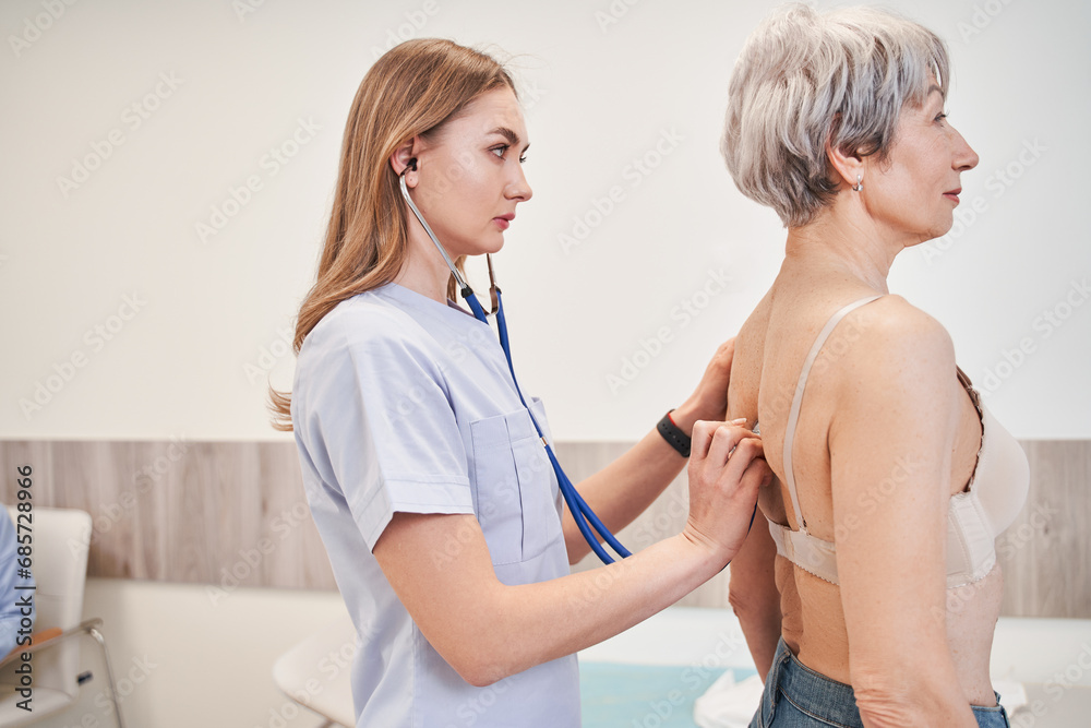Experienced practitioner standing behind the patient and carefully listening to her lungs Experienced practitioner standing behind the patient and carefully listening to her lungs
